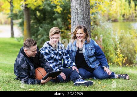 Ein junger Mann mit Down-Syndrom sitzt mit seiner Familie und spielt auf einem Tablet, während er sich in einem Stadtpark auf einem warmen ... Stockfoto