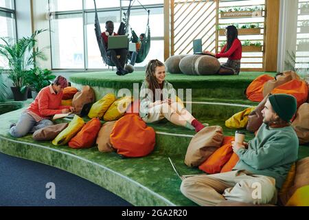 Mehrere junge interkulturelle Manager von großen modernen Open Space Büro Mit Laptops, während Sie auf grünem Boden gegen Kissen sitzen Stockfoto