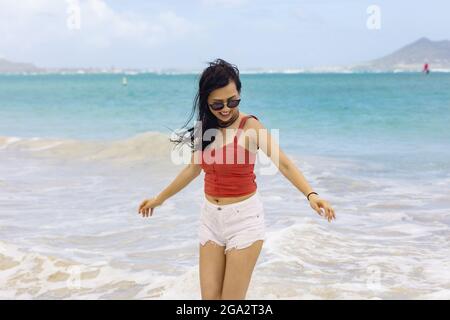 Eine Frau steht in der Brandung und schaut hinunter auf das Wasser, während sie einen tropischen Urlaub entlang der Küste von Oahu, Hawaii, genießt Stockfoto