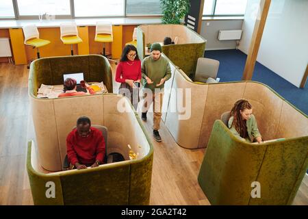 Mehrere junge interkulturelle Mitarbeiter in Casualwear sitzen an Schreibtischen in Büro und Blick auf Laptop-Displays während der individuellen Arbeit Stockfoto