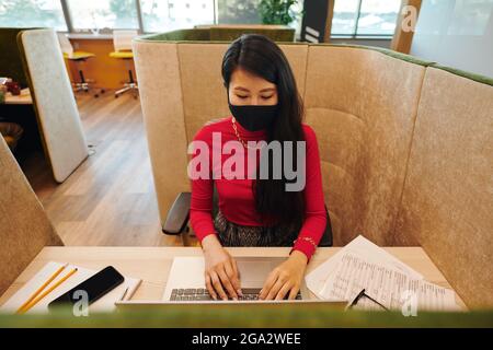 Asiatische junge Geschäftsfrau in smart Casualwear und Schutzmaske suchen Am Laptop-Display, während Sie am Schreibtisch sitzen und sich vernetzen Stockfoto
