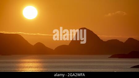 Mitternachtssonne, die hell am orangefarbenen Himmel über den Berggipfeln leuchtet und sich über den Nordatlantik reflektiert; Lofoten, Polarkreis, Norwegen Stockfoto