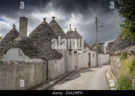 Straßenszene mit einer Reihe traditioneller apulischer runder Steinhäuser Trulli mit stürmischem Himmel an einem sonnigen Tag in der Stadt Alberobello Stockfoto