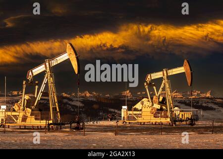 Pumpjacks, beleuchtet von goldenem Sonnenlicht vor einem dramatischen Sonnenuntergang am Himmel mit Wolken im Winter, westlich von Airdrie; Alberta, Kanada