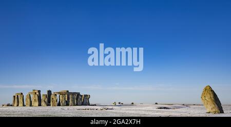 Stonehenge, definiert durch frühmorgendlichen Schnee mit einem strahlend blauen Himmel und dem schrägen Heel Stone rechts vom Steinkreis Stockfoto