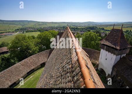 Nahaufnahme des gefliesten Dachs der Sächsischen Wehrkirche Viscri mit seiner umgebenden Steinmauer und einem Überblick über das Dorf und die Landschaft Stockfoto