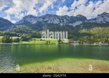 Spiegelung des grasbewachsenen Ufers rund um den Hintersee unter den bayerischen Alpen mit Unterkünften und Menschen, die das Wasser in Tretbooten genießen Stockfoto
