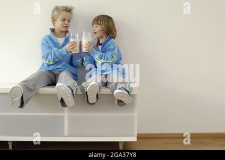 Zwei Jungen sitzen in passenden Outfits und toasten mit vollen Gläsern Milch; Deutschland Stockfoto