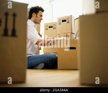 Ein Mann sitzt auf einem Laptop, der auf einer beweglichen Kiste aufgesetzt ist, umgeben von verpackten Kästen in einer häuslichen Umgebung; Deutschland Stockfoto