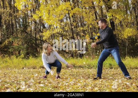 Ein reifes Ehepaar, das in der Herbstsaison gemeinsam in einem Stadtpark viel Zeit verbringt und Spaß dabei hat, sich gegenseitig heruntergefallene Blätter zu werfen Stockfoto