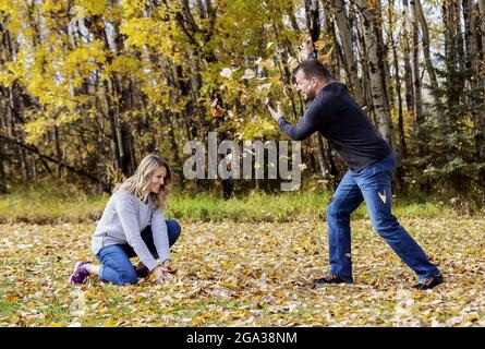 Ein reifes Ehepaar, das in der Herbstsaison gemeinsam in einem Stadtpark viel Zeit verbringt und Spaß dabei hat, sich gegenseitig heruntergefallene Blätter zu werfen Stockfoto