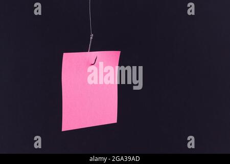 Nachbau eines leeren rosa Notizpapiers mit Kopierraum, das auf einem Angelhaken gegen den schwarzen Hintergrund aufgehängt wird. Erinnerung oder Aufgabenliste. Haftnotiz-Vorlage Stockfoto