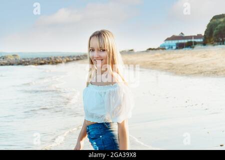 Porträt einer glücklichen lächelnden Frau in weißem Oberteil und Jeans-Shorts, die den Moment bei einem Spaziergang am Strand genießt. Sommerferien, Urlaub Stockfoto
