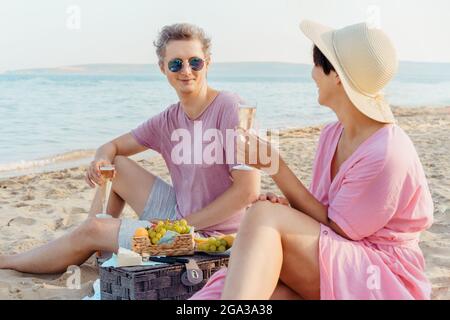 Lachendes Paar, das ein Picknick am Strand am Meer hat. Mann und Frau in der Liebe halten Gläser mit Wein oder Champagner, feiern den Moment mit Meer Stockfoto