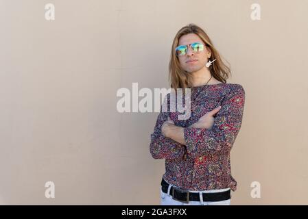 Porträt eines jungen Hipstermannes mit langen Haaren, der an einer beigen Wand mit gekreuzten Armen steht Stockfoto
