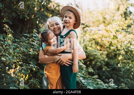 Großmutter mit zwei Enkelkindern, die im Garten stehen und die glückliche Familie umarmen und lachen. Stockfoto