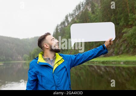 Der junge Mann mit Bart in der Jacke sagt alle Wörter mit der Möglichkeit, sie auf weißem rechteckigen Hintergrund zu schreiben, die er mit der Hand gegen b hält Stockfoto