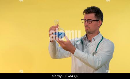 Nahaufnahme des jungen Arztes im Labormantel und Brillen, die mit blauer Flüssigkeit auf den Kolben blicken. Es ist ein gelber Hintergrund vorhanden. Stockfoto