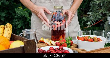 Lebensmittelbanner. Glasflasche mit erfrischender hausgemachter Limonade oder Sangria-Punsch mit Zitrusfrüchten und Bio-Beeren. Frau Hände halten einen Sommer-Cocktail Stockfoto