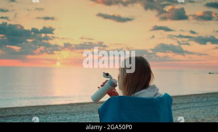Junge Frau trinkt aus Thermalbecher und sitzt am Strand vor Sonnenaufgang. Rückansicht. Stockfoto