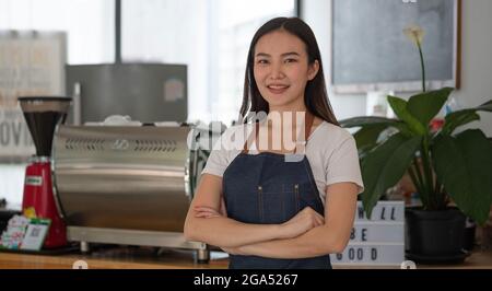 Schöne asiatische Frau Ladenbesitzer mit stehen in ihrem Café Blick auf Kamera und lächelnd.Portrait von Mädchen Kellnerin trägt Schürze und stehend Stockfoto