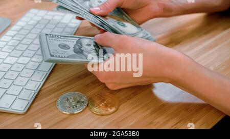 Schnittansicht der Hände von Frauen, die große Bündel von Dollarscheinen auf Holzschreibtisch mit Bitcoins und Tastatur zählen Stockfoto