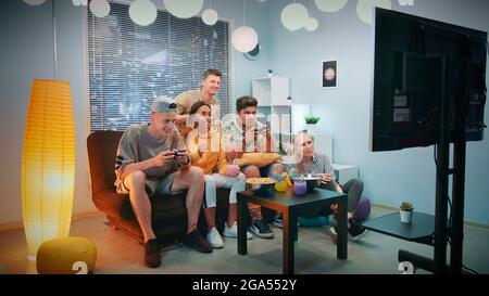 Zwei Spieler, die im Konsolenspiel spielen, einer davon gewinnt. Ihre Freunde jubeln sie hoch, sitzen auf dem Sofa zu Hause, essen Snacks und haben Spaß. Stockfoto