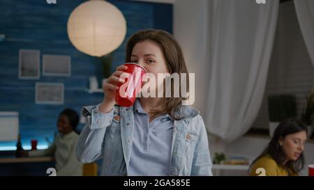 Nahaufnahme eines Frauenportraits, das in die Kamera schaut, während er im Wohnzimmer Bier trinkt, um den Geburtstag eines Freundes spät in der Nacht zu feiern. Im Hintergrund treffen sich multiethnische Freunde, die Spaß haben Stockfoto