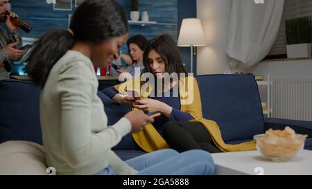 Eine Gruppe gemischter Renner, die spät in der Nacht im Wohnzimmer zusammenhängen und online Nachrichten auf dem Handy suchen. Multirassische Freunde, die während einer Überraschungsparty einen Unterhaltungsfilm ansehen Stockfoto
