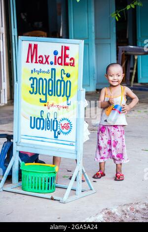 Das junge burmesische Mädchen mit rasiertem Kopf und Schlafanzug steht neben der Werbetafel in der Straße, Monywa, Myanmar Stockfoto