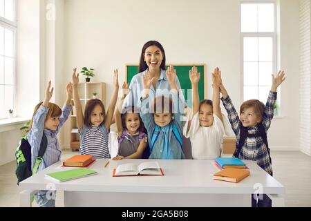 Fröhliche Lehrerin und ihre lustigen Grundschüler stehen mit erhobenen Armen. Stockfoto