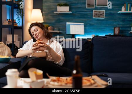 Frau, die mit Freunden telefoniert, während sie einen leckeren leckeren Burger in den Händen hält und sich spät in der Nacht auf der Couch im Wohnzimmer entspannt. Kaukasische Frau genießt Tajeaway Junk Food nach Hause geliefert Stockfoto