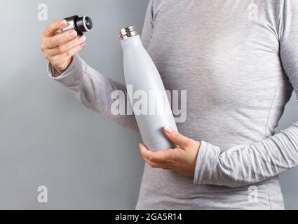 Nahaufnahme einer Frau im grauen T-Shirt mit einer grauen Isolierflasche auf grauem Hintergrund Stockfoto