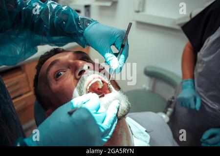 Kaukasischer männlicher Klient sitzt auf einem Zahnarztstuhl, während die Krankenschwester an den Zähnen operiert Stockfoto