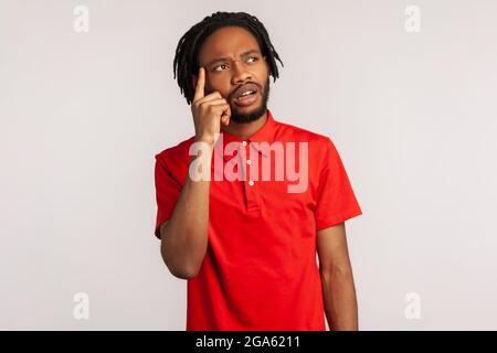 Porträt eines jungen attraktiven bärtigen Mannes mit Dreadlocks in einem roten T-Shirt im Casual-Stil, der denkt, hat eine neue Idee, sieht mit nachdenklichen Ausdruck aus Stockfoto