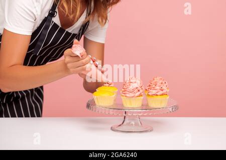 Zugeschnittenes Bild einer Frau, die Muffins mit rosiger Creme mit Konditor, köstlichem Dessert auf Glasplatte, gesichtsloser Konditorei schmückt. I Stockfoto