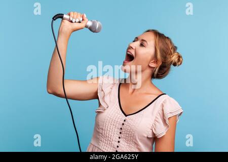 Seitenansicht positive blonde Frau im Sommerkleid laut singen Lied, hält Mikrofon in der Hand, Spaß beim Ausruhen in Karaoke, Sänger. Studio im Innenbereich Stockfoto