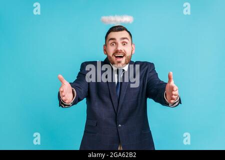 Kommen Sie hierher. Ein hübscher, freundlicher Mann im offiziellen Business-Anzug mit Nimbus über dem Kopf, der sich als Engel ausgibt und die Hände bereit hält, sich zu umarmen. Indoor st Stockfoto