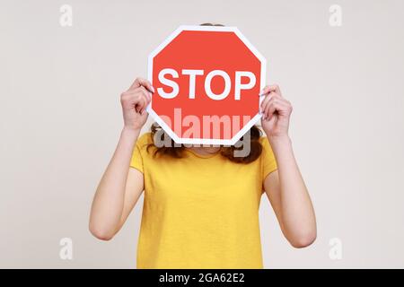 Weg verboten! Portrait eines anonymen Mädchens in gelbem Casual T-Shirt, das Gesicht mit Stoppsymbol bedeckt, rotes Verkehrsschild in der Hand, unzulässiger Zugang. Innen Stockfoto