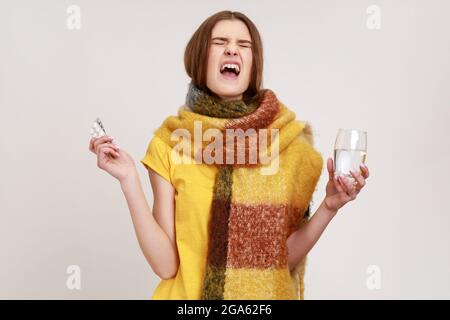 Porträt eines grippekranken Teenagers, das in einen Schal gewickelt ist und Pillen und ein Glas Wasser hält, vor Hass schreiend, sich über die Influenza-Krankheit genervt fühlt Stockfoto