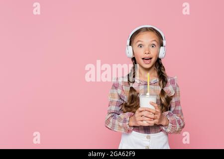 Aufgeregtes Kind in Kopfhörern, das Milchshake isoliert auf Rosa hält Stockfoto