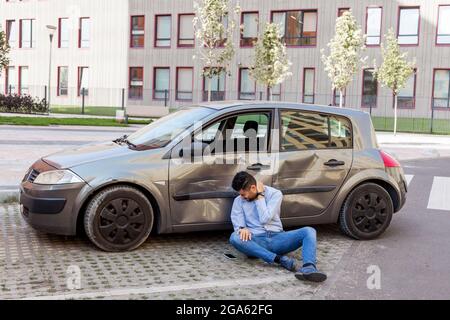 Mann in Jeans und Hemd mit Verletzungen sitzt auf dem Boden in der Nähe von gebrochenem Auto und den Hals berühren, fühlt schreckliche Schmerzen, Autounfall verletzt männlich und Stockfoto