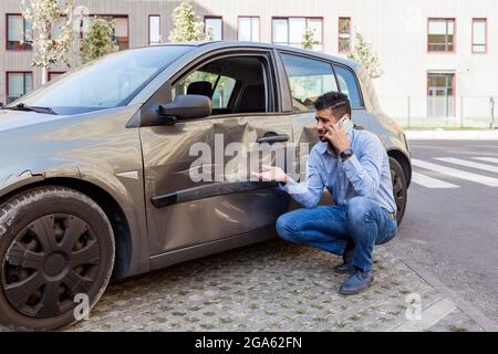 Hübscher bärtiger Mann in Jeans und blauem Hemd mit trauriger Gesichtsausdruck, telefoniert, ruft zur Polizei- oder Versicherungsinspektion, beschädigte Autotür mit Stockfoto