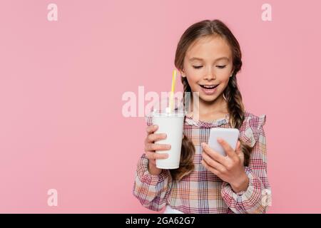 Lächelndes Kind mit Smartphone und hält Milchshake isoliert auf rosa Stockfoto