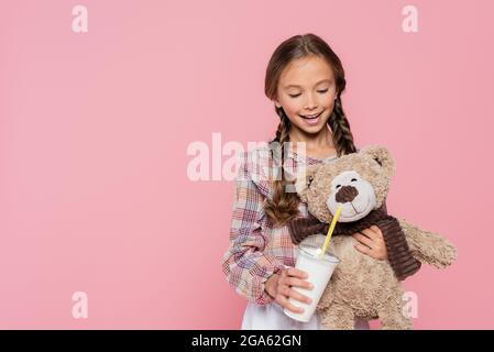 Kind im karierten Hemd, das Milchshake in der Nähe des Teddybären hält, isoliert auf Rosa Stockfoto