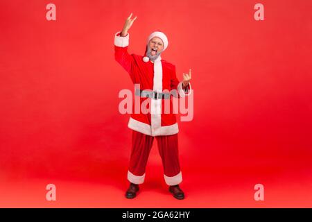 In voller Länge aufgenommen mit einem aufgeregten älteren Mann mit grauem Bart, der das weihnachtsmann-Kostüm trägt und Rock and Roll vor der Kamera singt und mit einem verrückten Look schreit Stockfoto