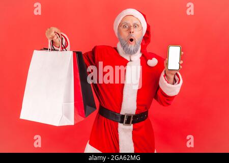 Überraschte ältere Mann trägt weihnachtsmann Kostüm mit Einkaufstaschen und Telefon mit leerem Display, schockiert über Weihnachtsrabatte, Online-Kauf. I Stockfoto