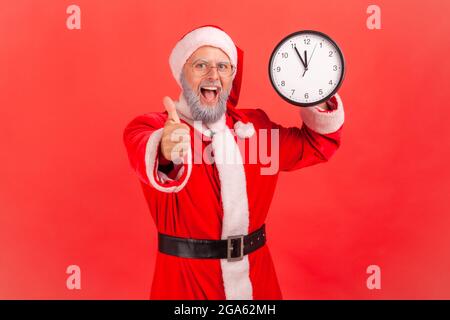 Glücklich aufgeregt älteren Mann mit grauem Bart trägt weihnachtsmann Kostüm zeigt Wanduhr und zeigt Finger zur Kamera, hat erstaunt Ausdruck. Innen Stockfoto