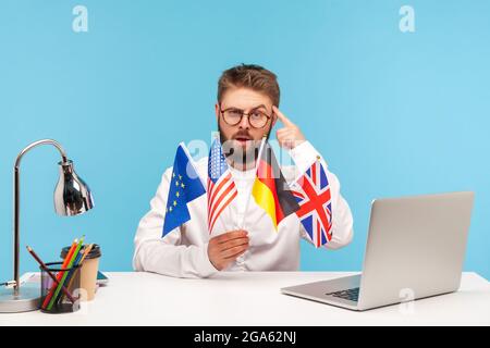 Intelligenter, seriöser Lehrer in Brillen, der Flaggen verschiedener Länder mit dem Finger auf den Kopf zeigt und vorschlägt, mit ihm Fremdsprache zu lernen. I Stockfoto