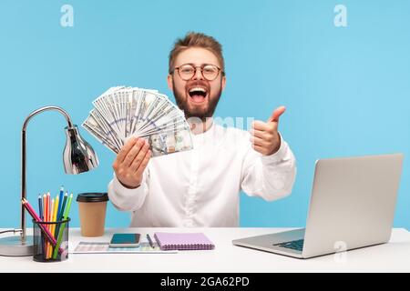 Glücklich aufgeregt bärtigen Mann zeigt Daumen nach oben wie Geste hält Fan von Dollar-Bargeld sitzen am Arbeitsplatz mit Laptop, easy Profit, illegale Gewinne. I Stockfoto